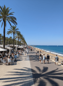Zomerse mediterrane boulevard met palmbomen, terrasjes en mensen die langs de kustpromenade wandelen bij helder weer